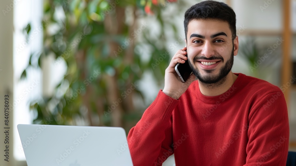 Poster smiling man in red sweater talking on smartphone while sitting at desk with laptop. casual and frien