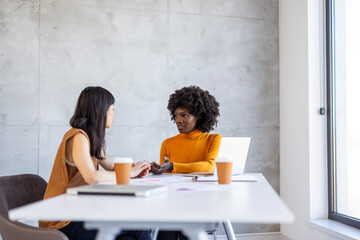 Black and Asian business professionals engage in discussion, with a laptop and coffee on the table, in a bright contemporary office setting.