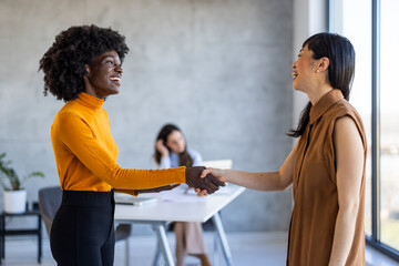 A professional Asian businesswoman and her counterpart with African ethnicity greet each other with a handshake in a sleek, modern office environment, exemplifying teamwork and partnership.