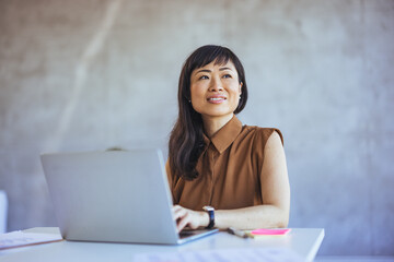 A cheerful Asian businesswoman engages with her work in an office, smartly dressed in professional attire, surrounded by office supplies and utilizing a laptop.