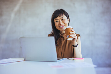 A joyful Asian businesswoman enjoys a coffee break at her office space, equipped with a laptop and...