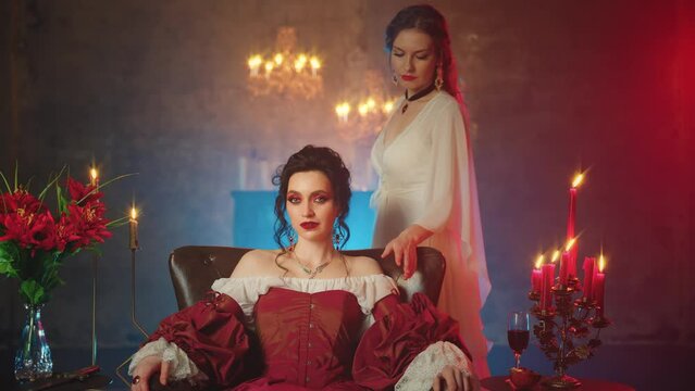 Two women vampires. beauty woman old style black hair Fantasy lady evil queen sitting on throne. Evil vampire girl sexy face red lips eyes long dress with friend maiden old style. dark night room art