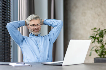 Mature businessman enjoying a break at home office desk, smiling, and looking relaxed with hands...