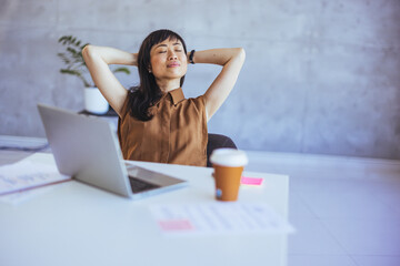 A serene Asian businesswoman leans back with eyes closed, enjoying a break in a well-lit office,...