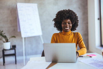 An enthusiastic Black businesswoman engages with her laptop at a bright office desk, donning...
