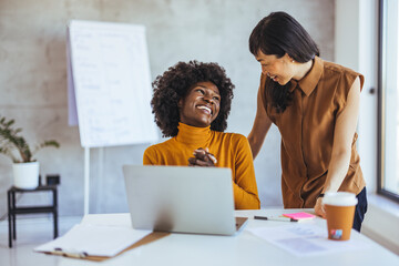 An Asian businesswoman pairs with a Black colleague, sharing insights over a laptop in a bright...
