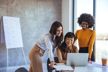 Three women of different ethnicities smiling and working together at a laptop in a bright, stylish...