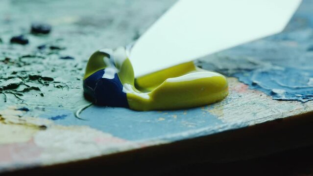 Painting knife mixing yellow and blue oil paints squeezed on canvas while creating new color. Extreme close-up view