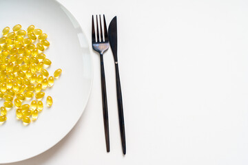 Lots of clear yellow fish oil pills, omega 3 on a white plate with fork and knife on a light background. Close-up. Copy space