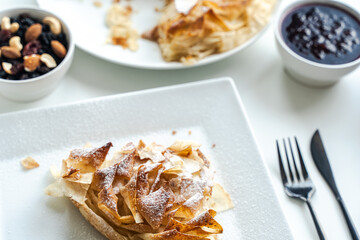 A slice of apple pie or a dessert of phyllo pastry on a white plate on a light background with nuts and jam. Selected focus.