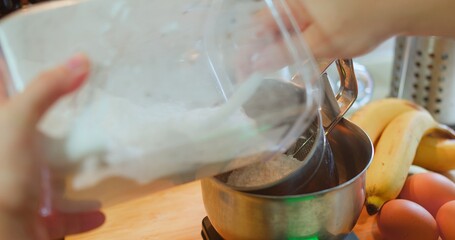 Close-up of a person pouring flour into a stainless steel mixing bowl on a kitchen scale, surrounded by fresh ingredients like bananas and eggs, capturing the process of baking or cooking at home