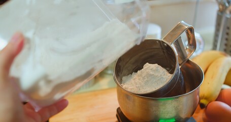 Close-up of a person pouring flour into a stainless steel mixing bowl on a kitchen scale, surrounded by fresh ingredients like bananas and eggs, capturing the process of baking or cooking at home