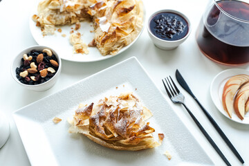 A slice of apple pie or a dessert of phyllo pastry on a white plate on a light background with nuts and jam. Selected focus.
