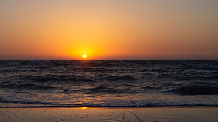 Obraz premium Beautiful sunset over a sandy beach with the sun setting into the sea. Golden light reflects off the calm waves, creating a serene and picturesque coastal scene perfect for relaxation and nature-theme