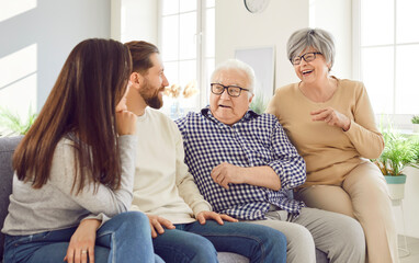 Happy Caucasian elderly parents and adult children sit together on the sofa at home, talking and...