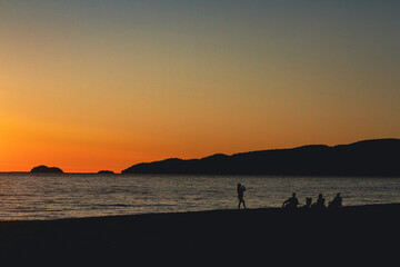 Obraz premium While watching the evening sunset on the shores of Agawa Bay campground a silhouetted young woman takes a photograph of her family while they all relax along the beach