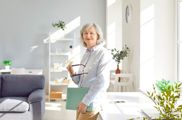 Portrait of confident smiling senior business woman standing in living room at home and looking...