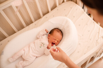 Mother give pacifier to baby girl in crib at home close up top view. Motherhood.