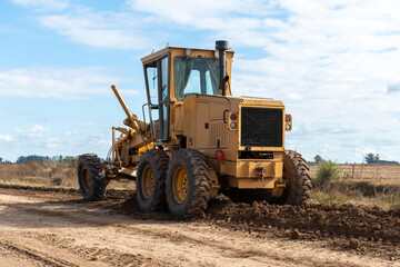 Heavy machinery is working on a dirt road in a rural setting under a partly cloudy sky. The image showcases two yellow graders and a green tractor, emphasizing construction and agricultural themes