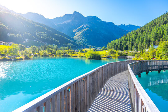 A tranquil wooden boardwalk bridge curves above the vibrant blue waters of Lake in Sulden, Italy. With the majestic Italian Alps towering in the background under a clear azure sky.