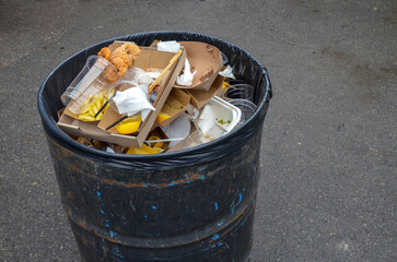 Overflowing black trash bin outdoors, filled with various types of garbage including food...