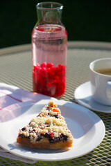 A piece of homemade cake with red and black currant and crumb on a white plate, a cup of black coffee with saucer, currant lemonade and a striped tea towel on a white garden table. Summer still life.