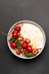 Greek yogurt with granola and fresh strawberries, cherry and honey, decorated with aromatic mint leaves on black stone background top view. Healthy tasty homemade breakfast concept. Vertical image.