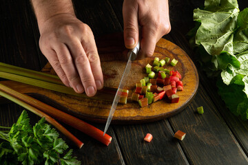 A chef uses a knife to cut rhubarb on a wooden cutting board before preparing a vitamin salad for dinner. Vegetarian cooking concept.