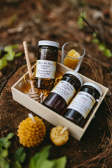 Gift set of jars with various organic honeys in a white box and candles made of natural beeswax in the shape of flowers and pine cones on an old stump in forest. Artistic photo of beekeeping products