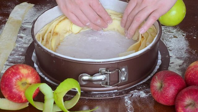Assembling apple pie into springform pan. Women's hands place thin strips dough slices apples spiral pie in shape of rose. Tea treat with fruits in foreground, red green peel. Cozy homemade baking, 4K