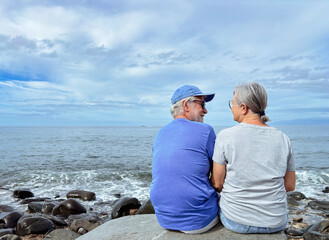Rear view of bonding family senior couple sitting on a pebble beach at sea looking each other face...