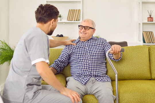 Portrait of a young nurse sitting back talking with a senior patient with crutches sitting on the sofa at home. Physiotherapist helping old man in rehab. Recovery and nursering home concept.