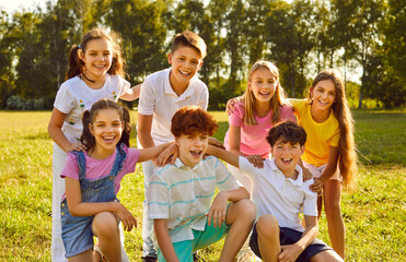 Fototapeta premium Portrait of a happy smiling children friends sitting on the green grass together in the park outdoors and looking cheerful at the camera hugging and having fun. Kids summer camp and holidays concept.