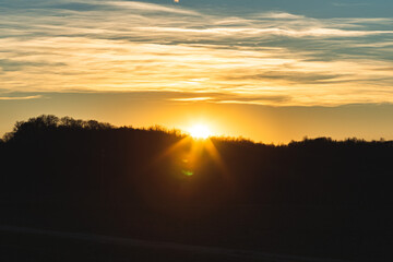 sole dalle sfumature di colore giallo e arancione, visto in controluce, che tramonta dietro la sagoma di una collina e con un cielo parzialmente sereno e coperto da nuvole leggere