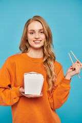 A beautiful woman with blonde hair delicately holds food box and chopsticks, savoring Asian cuisine.