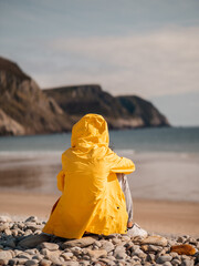 A person is sitting on a beach in a yellow raincoat. The beach is rocky and the ocean is in the...