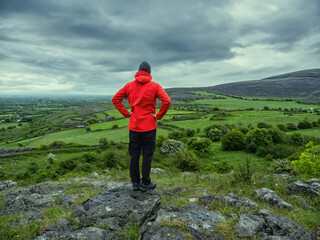 Obraz premium Man in red jacket looking at stunning nature scenery with mountains and dramatic sky. Burren, Ireland. Irish nature scene. Travel and tourism concept. Outdoor activity and sightseeing.