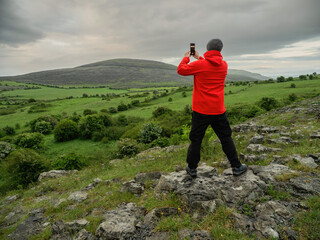 Obraz premium A male tourist in red outdoor jacket taking picture of stunning nature scene on his smart phone. Travel concept. Beautiful nature of Burren with hills and dramatic sky. West of Ireland.