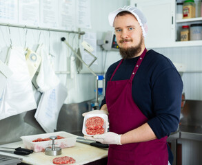 Positive young bearded butcher standing in meat processing room of local shop, offering freshly...