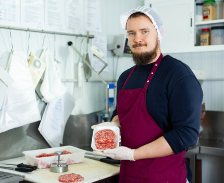 Positive young bearded butcher standing in meat processing room of local shop, offering freshly formed ground veal burger patties in plastic tray prepared for sale