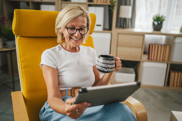 Mature senior woman sit at home hold cup of hot drink and use tablet