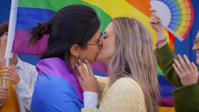 Young lesbian couple kissing on gay pride day outdoors. Group of LGBT community celebrating party on background with rainbow flag and fans. Types of sexuality on generation z