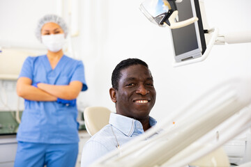 Obraz premium African-american man patient sitting on dental chair and smiling. Asian woman dentist standing in background.