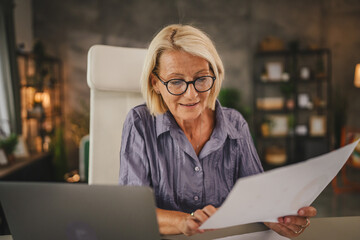 Mature senior woman work from home use laptop and hold documents