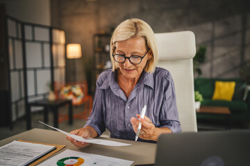 Mature senior woman work from home use laptop and hold documents
