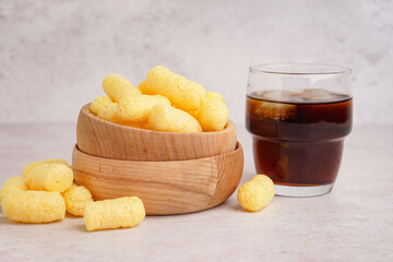 Wooden bowl with tasty salty corn sticks and glass of cola on light background, closeup