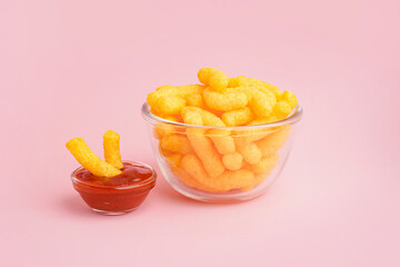 Glass bowl with tasty salty corn sticks and ketchup on pink background