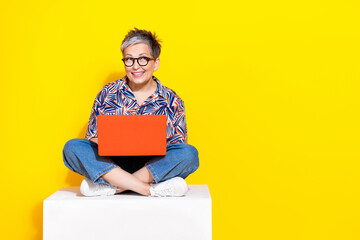 Full body photo of attractive retired woman sit cube hold netbook dressed stylish colorful clothes isolated on yellow color background