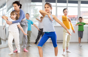 Fototapeta premium During dance workshop, girl with team of young like-minded children learn to dance modern dances. Woman teacher corrects movements of hands and body, instructs students to perform dance movements