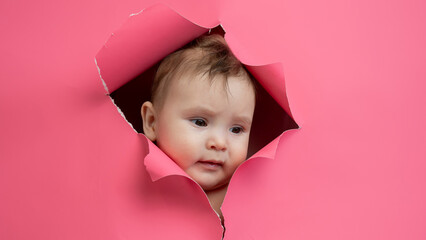 Cute Caucasian newborn baby boy peeks out of a hole in a paper pink background.